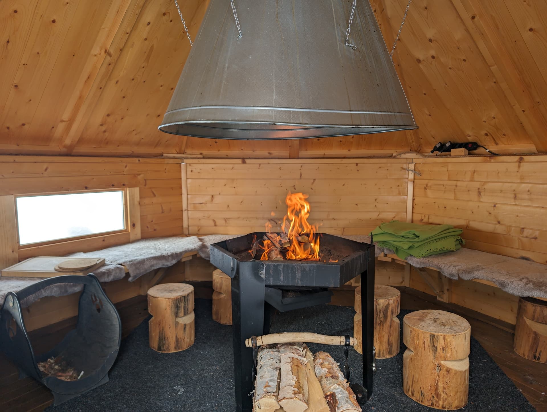 Blick in eine leere, holzverkleidete Kota mit zentraler Feuerstelle, Rentierfellbänken, Baumstumpf-Hockern und gestapeltem Birkenholz.