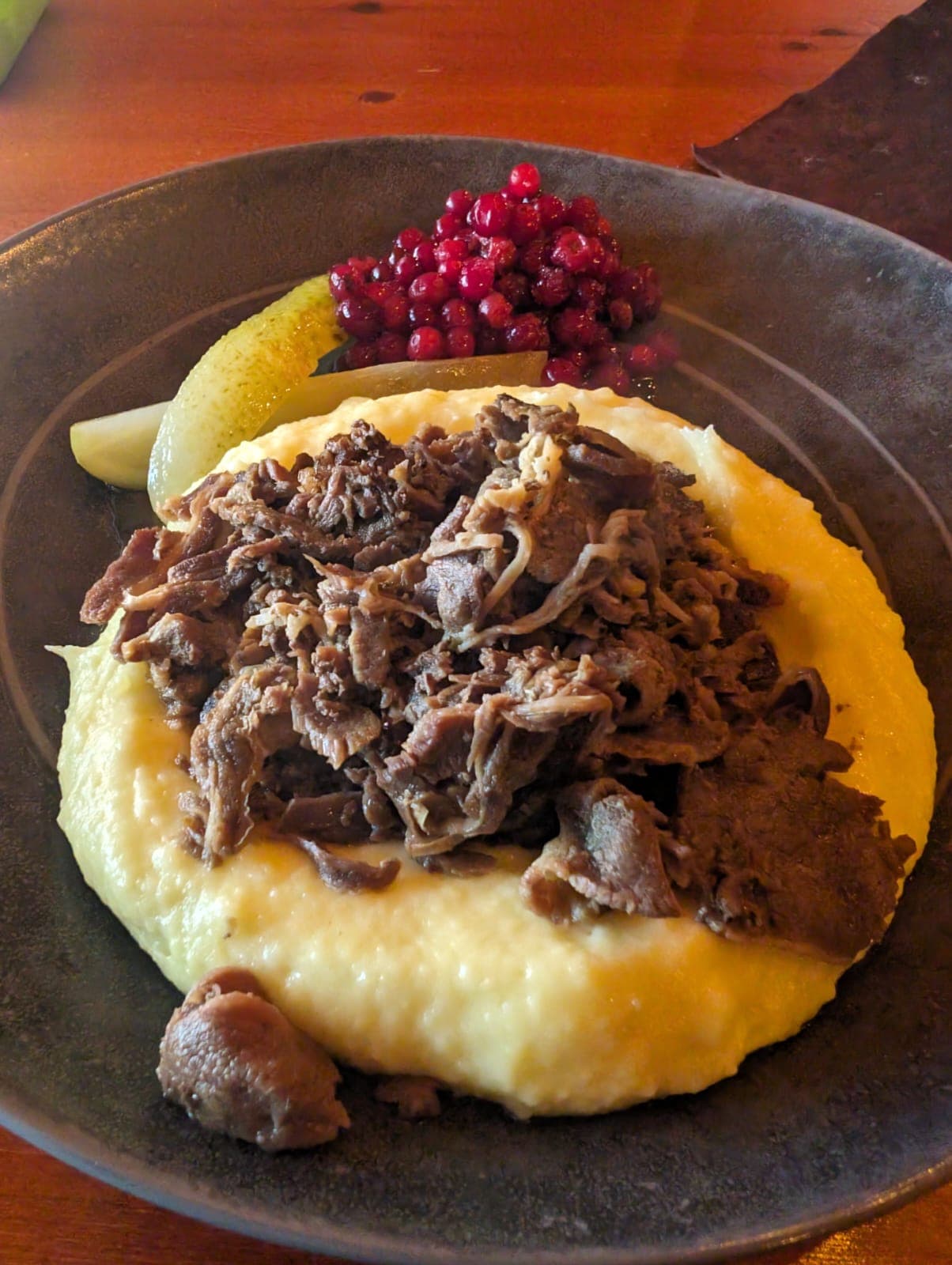 Auf einem dunklen Teller liegt lappländisches Rentierfleisch auf cremigem Kartoffelpüree, garniert mit Preiselbeeren und eingelegter Gurke.