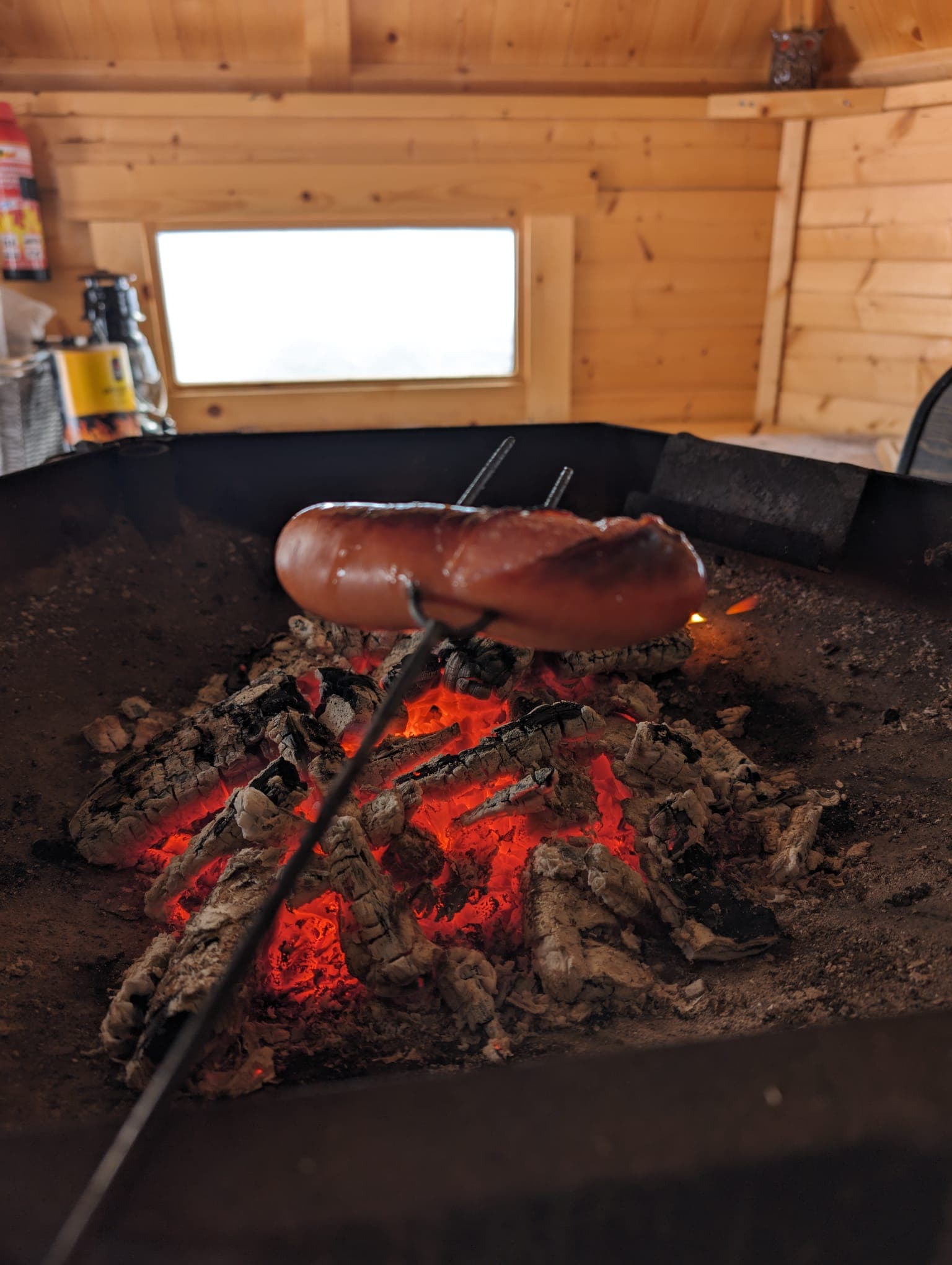 Eine Bratwurst am Stock röstet über der Glut eines offenen Feuers in einer traditionellen finnischen Grillhütte.