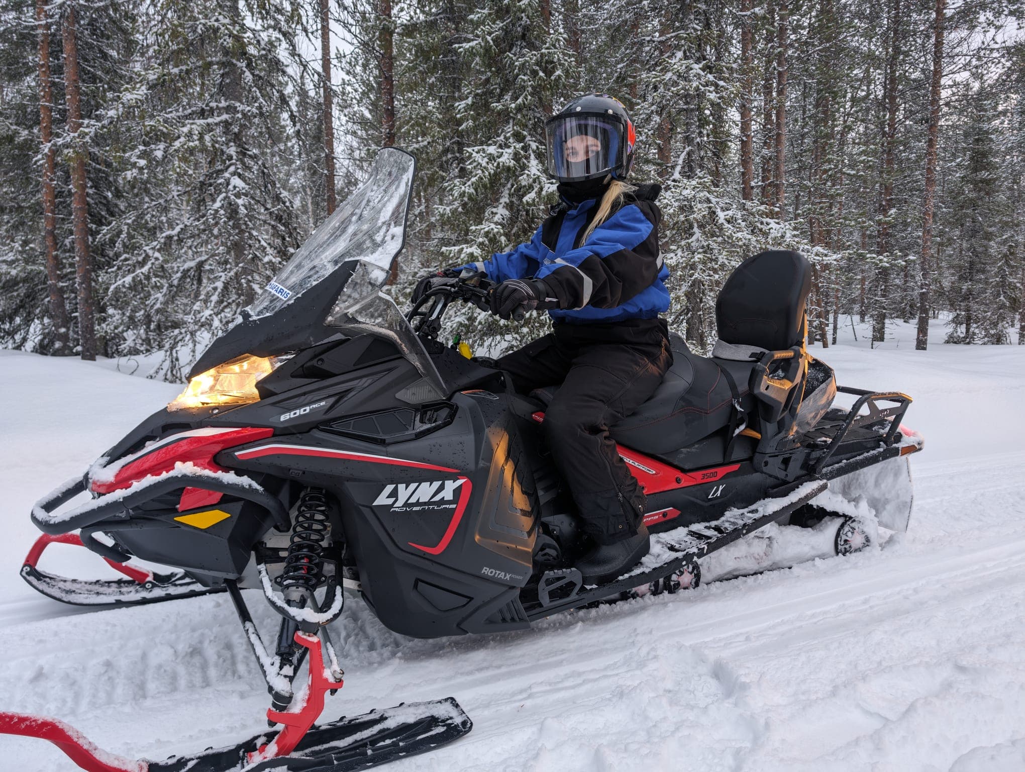 Eine Frau in Helm und Schneemobilkleidung sitzt auf einem schwarz-roten Schneemobil vor verschneiten Kiefern in Lappland.