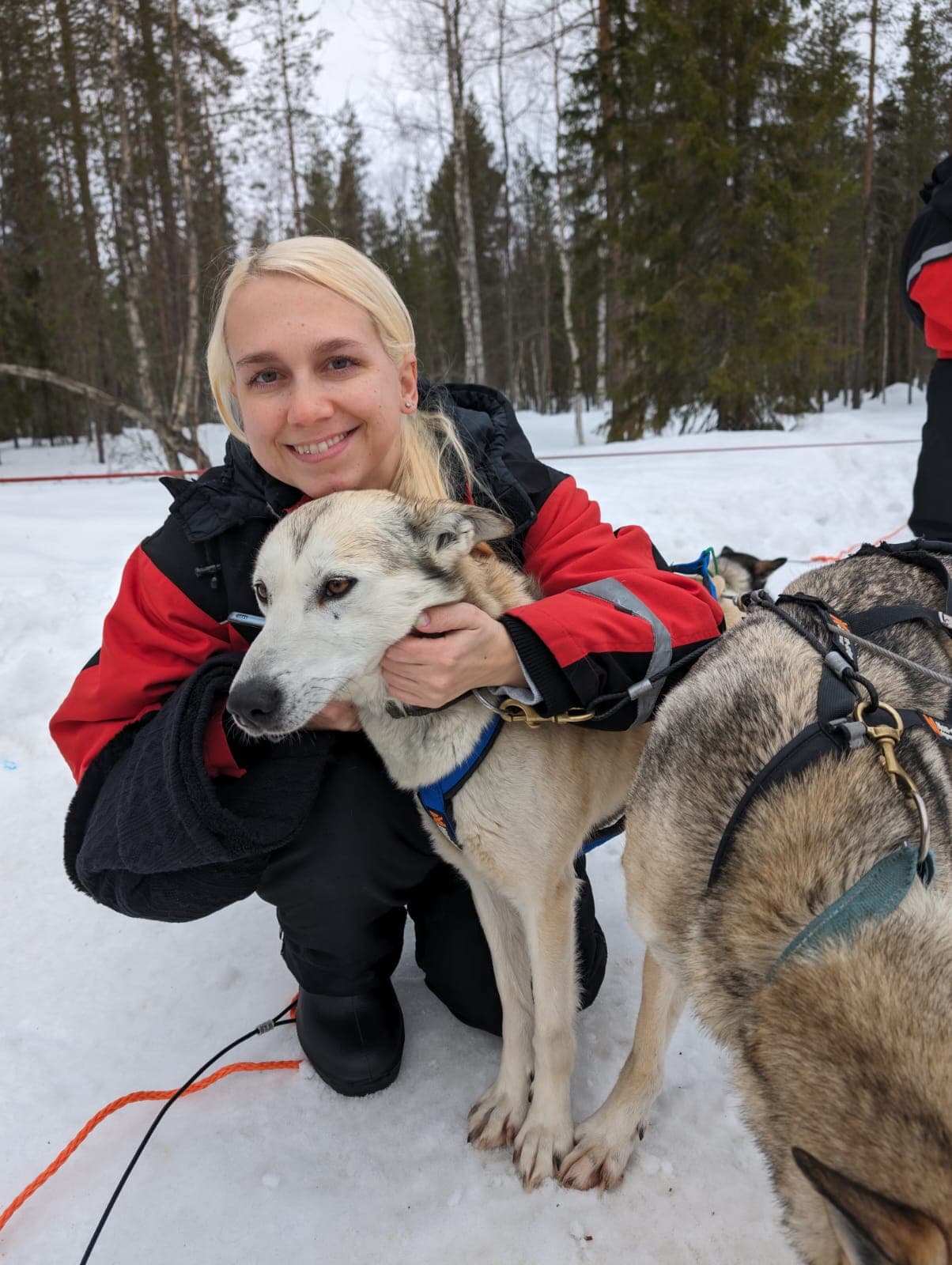 Eine Frau in roter Skijacke kniet im Schnee und umarmt den Kopf eines Schlittenhundes vor verschneiter Kiefernwald-Kulisse.