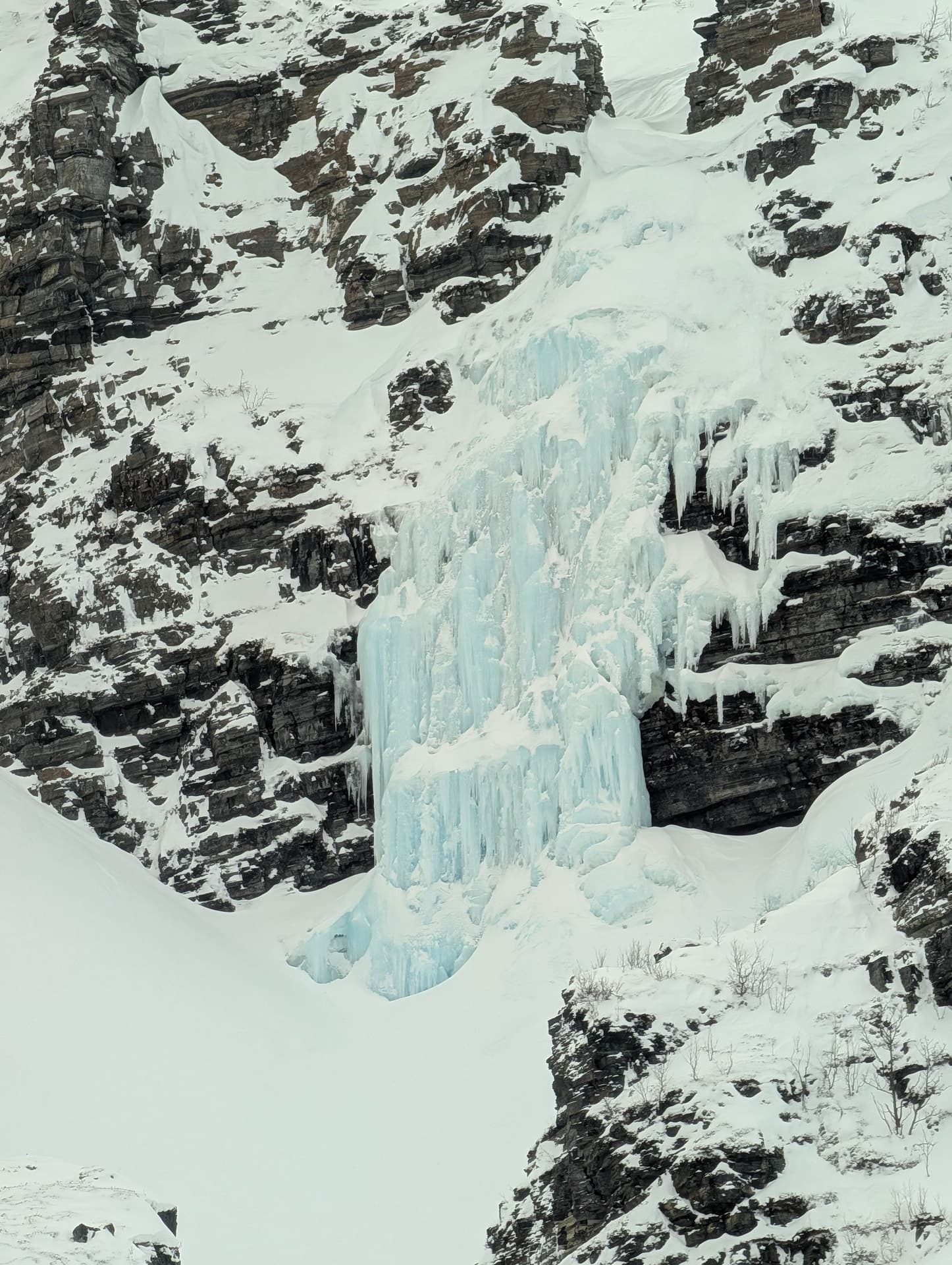 Eine steile Felswand wird von einer massiven Eissäule durchbrochen, die sich aus dem Schnee herauskristallisiert.