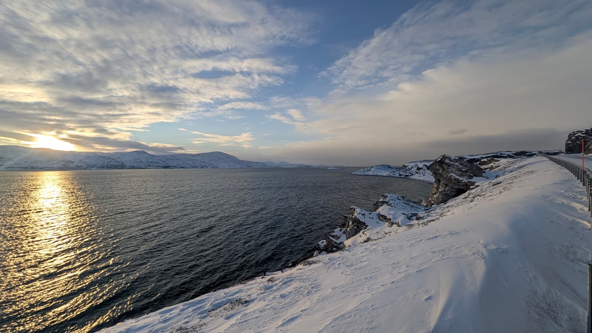 Eine ruhige Winterlandschaft an der norwegischen Küste bei tiefstehender Sonne, die sich auf dem dunklen Wasser spiegelt. Der schneebedeckte Pfad und die fernen Berge vermitteln eine friedliche, kalte Atmosphäre.