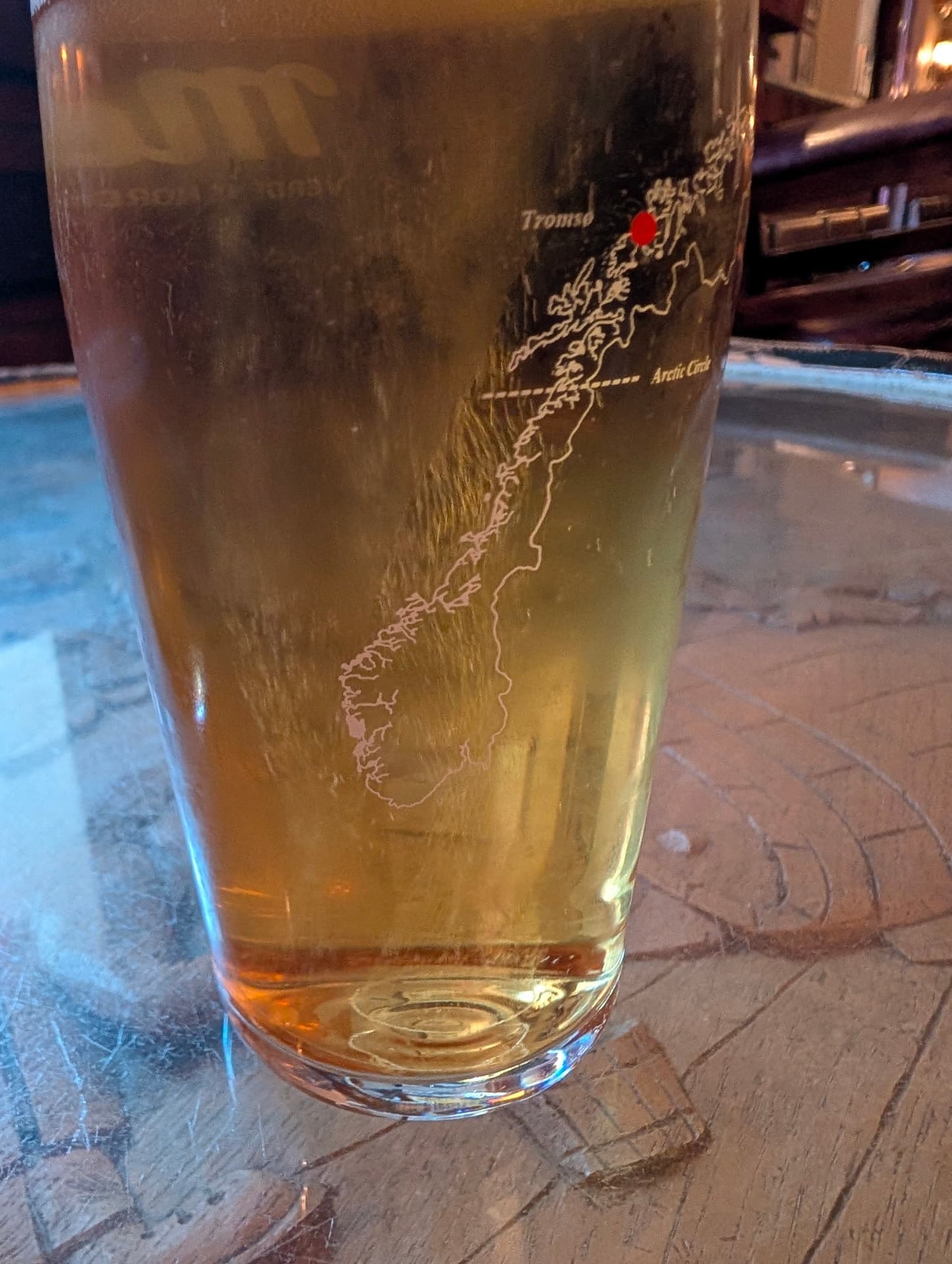 Ein Glas Bier in Ølhallen mit eingravierter norwegischer Küste, Tromsø ist rot markiert, auf einem rustikalen Holztisch.
