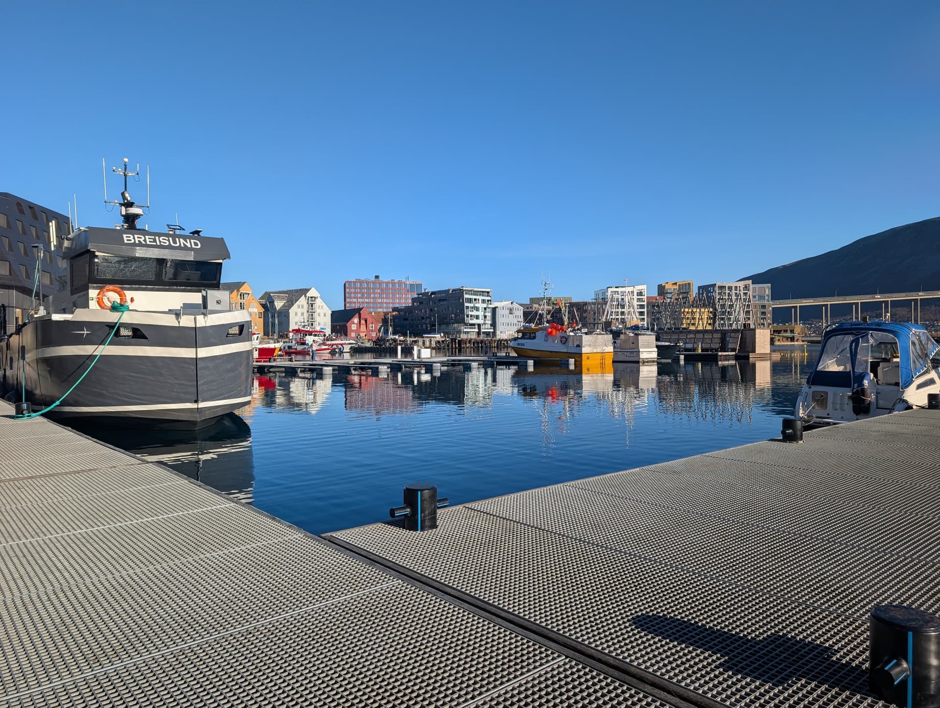 Ein graues Fischerboot mit dem Schriftzug 'BREISUND' liegt im ruhigen Hafen von Tromsø vor bunten Gebäuden und einem Berg unter blauem Himmel.