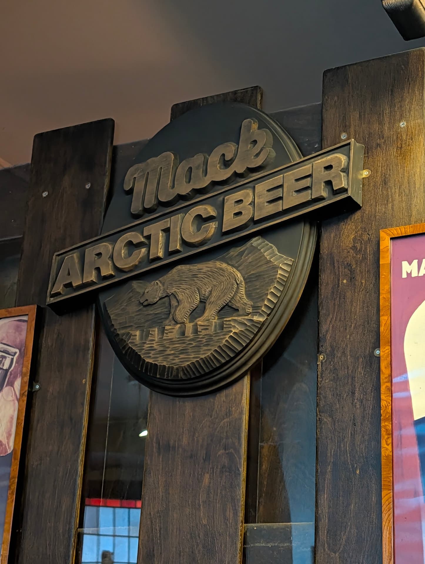 Ein geschnitztes Holzschild mit der Aufschrift 'Mack Arctic Beer' und einem Eisbären an der dunklen Holzwand von Ølhallen in Tromsø.