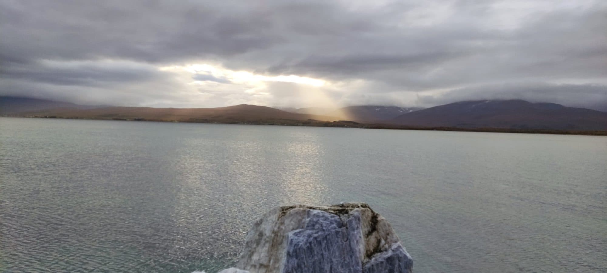 Blick über einen Fjord bei Tromsø auf herbstlich braune Berghänge, durch tiefe Wolken bricht Sonnenlicht, im Vordergrund ein Felsblock im Wasser.