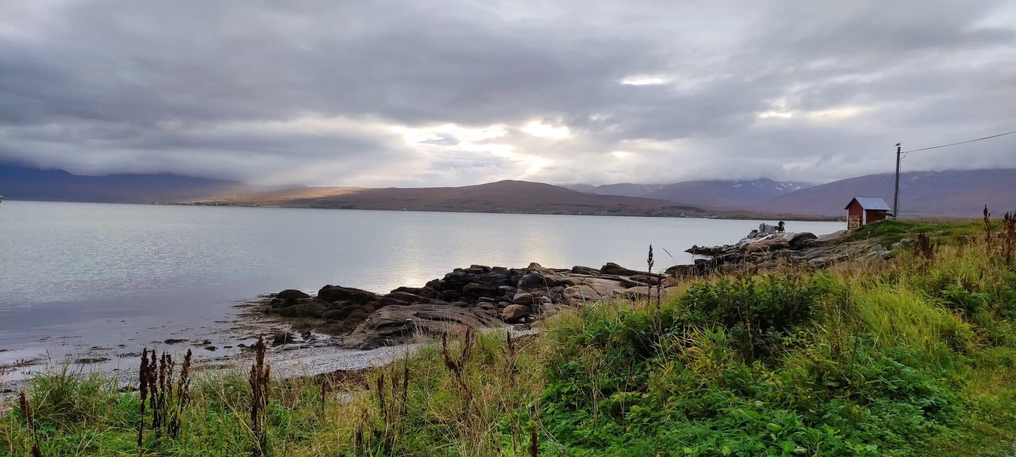Felsiges Ufer am Fjord bei Tromsø mit Gras und Steinen, am rechten Bildrand ein rotes Bootshaus, dahinter Berge unter Wolken.
