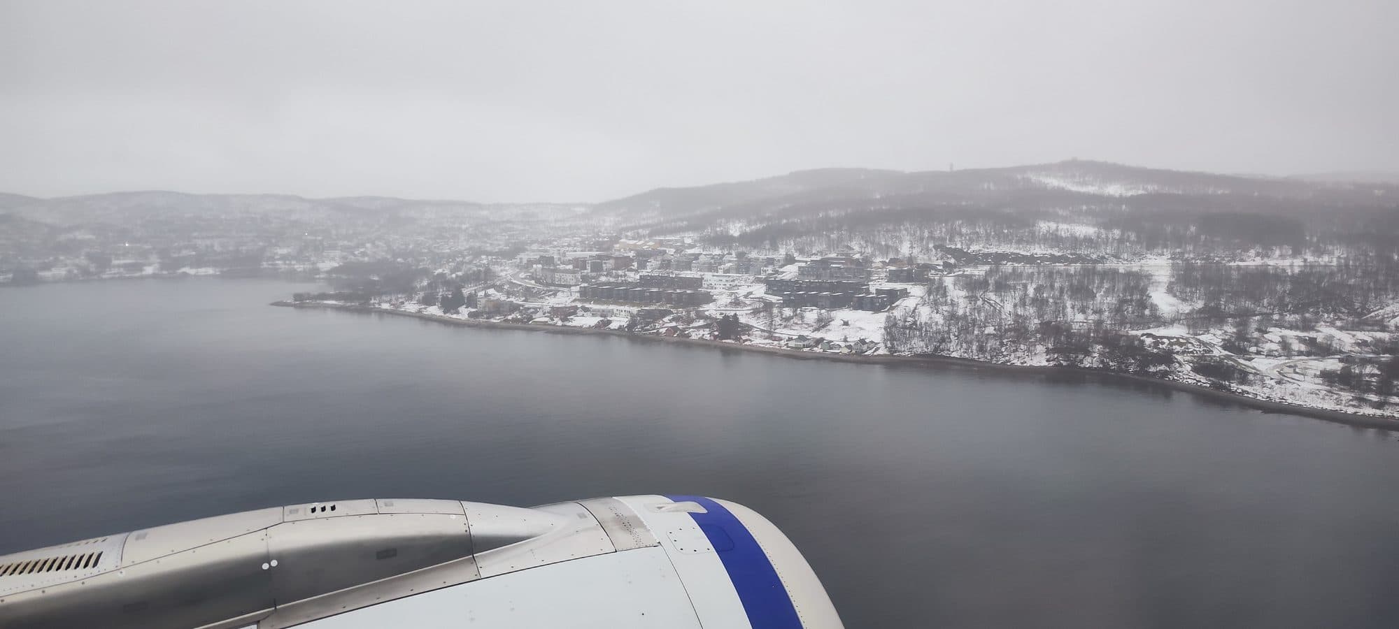 Blick aus dem Flugzeugfenster kurz vor der Landung auf eine verschneite Küstenstadt am Fjord, halb verhüllt von tiefen Wolken.