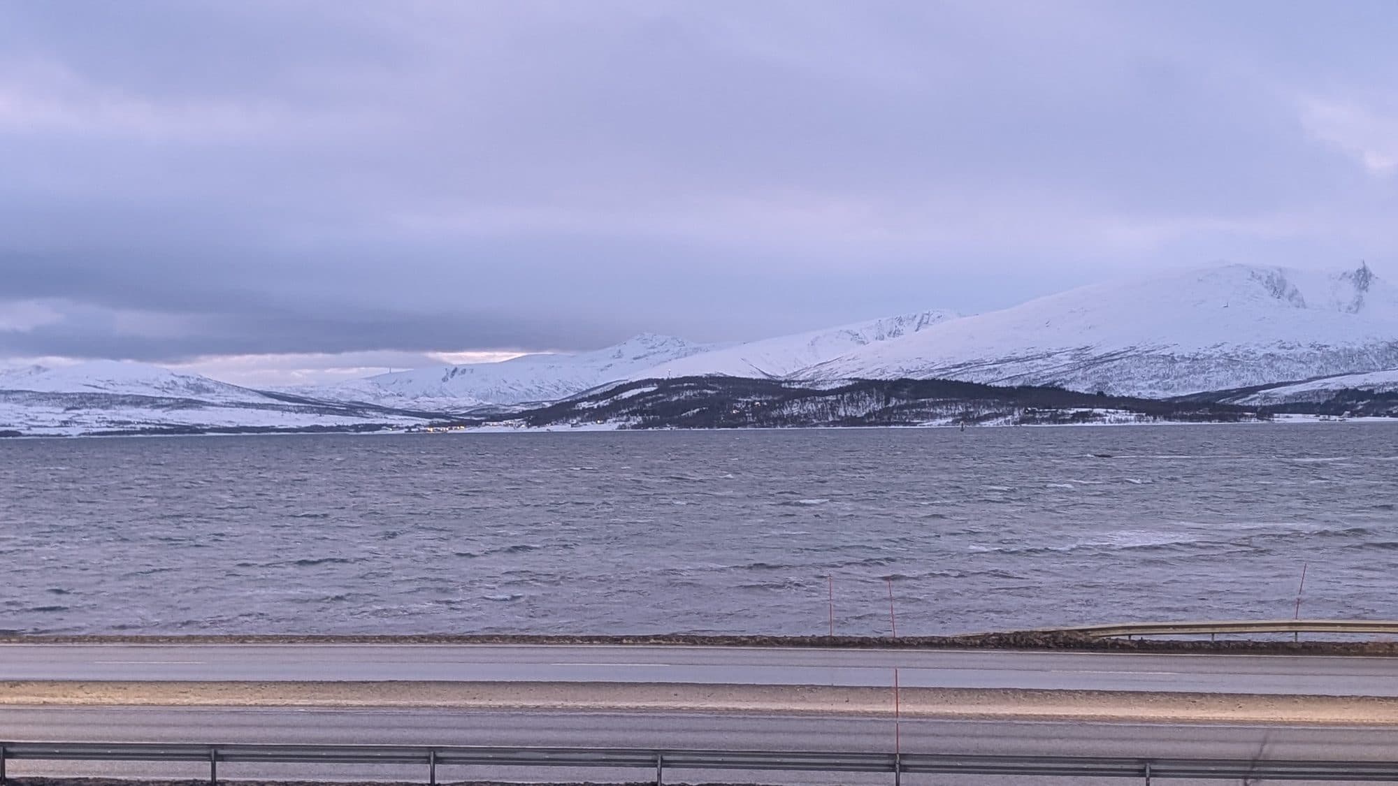 Weiter Blick über einen aufgewühlten Fjord bei Tromsø auf schneebedeckte Berggipfel unter dramatischem Wolkenhimmel.