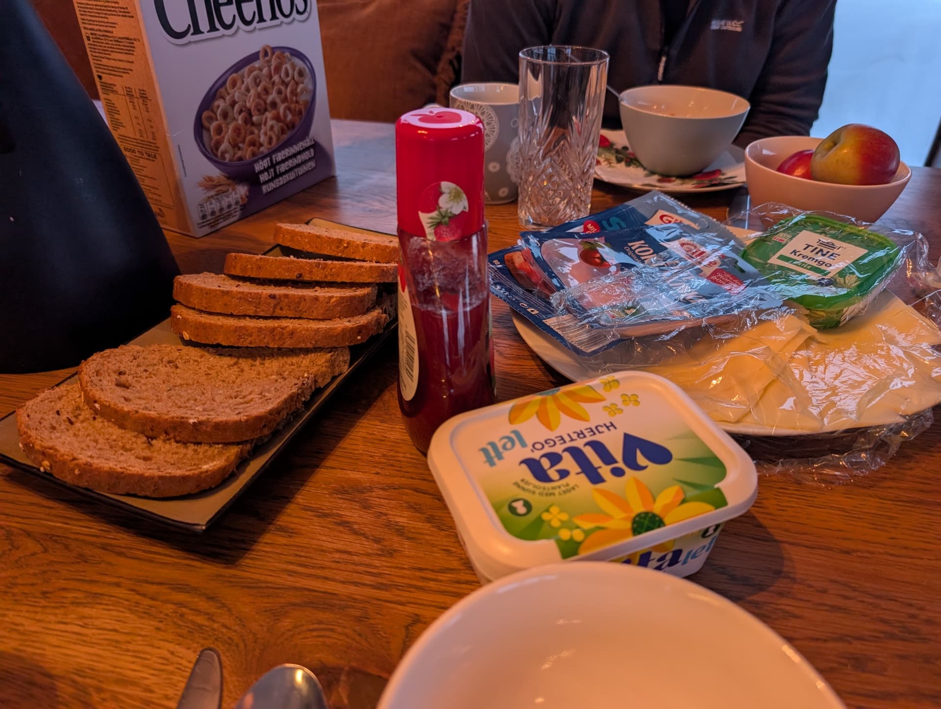 Ein rustikales Frühstück auf einem Holztisch mit typischen norwegischen Marken wie Vita-Butter und Tine-Käse, das einen authentischen Einblick in den Alltag im Ferienhaus bietet.